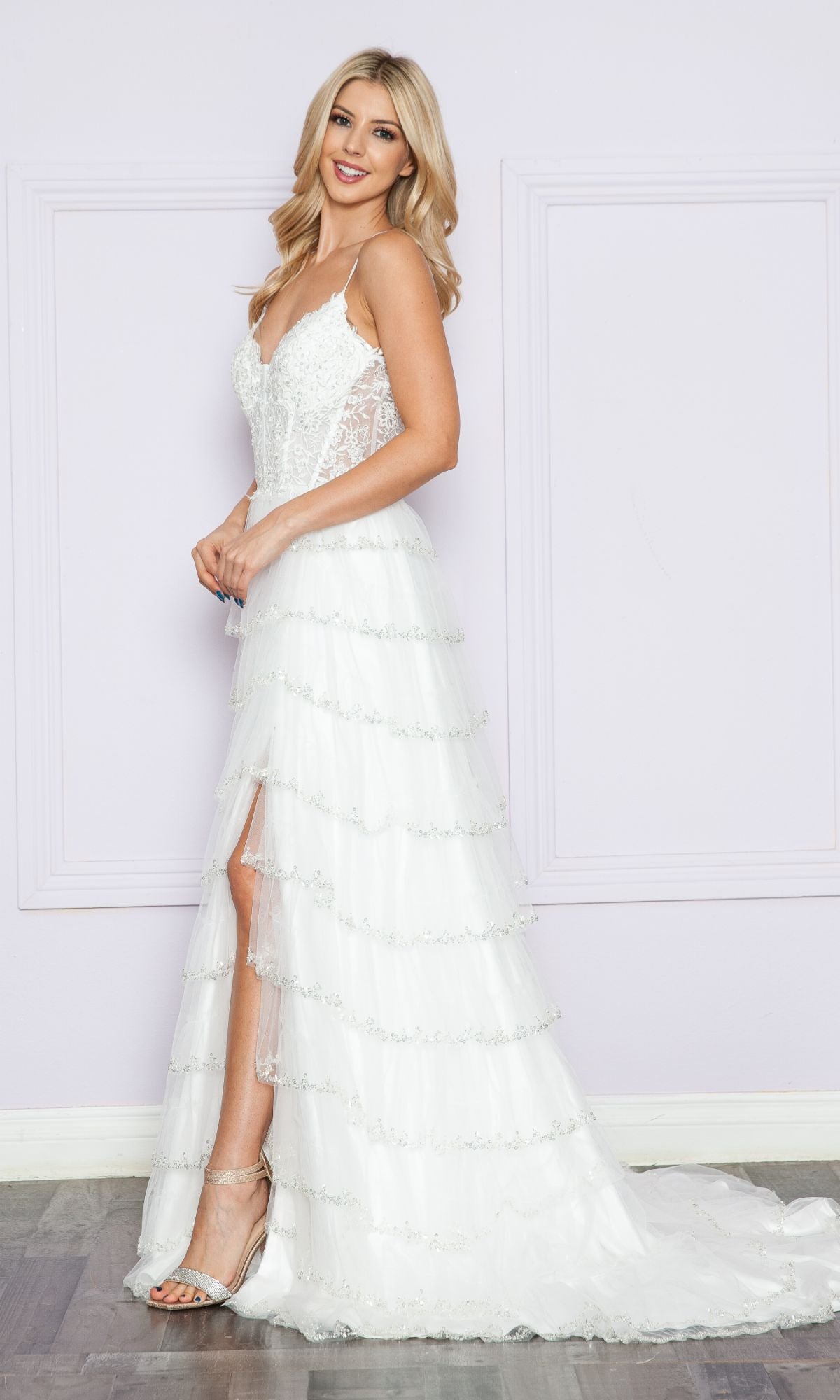 A woman stands indoors against a light purple wall wearing the Ruffled Long A-Line Tiered Prom Dress: Poly 9404, a white sleeveless gown with an embroidered V-neck bodice and layered skirt featuring a thigh-high slit.
